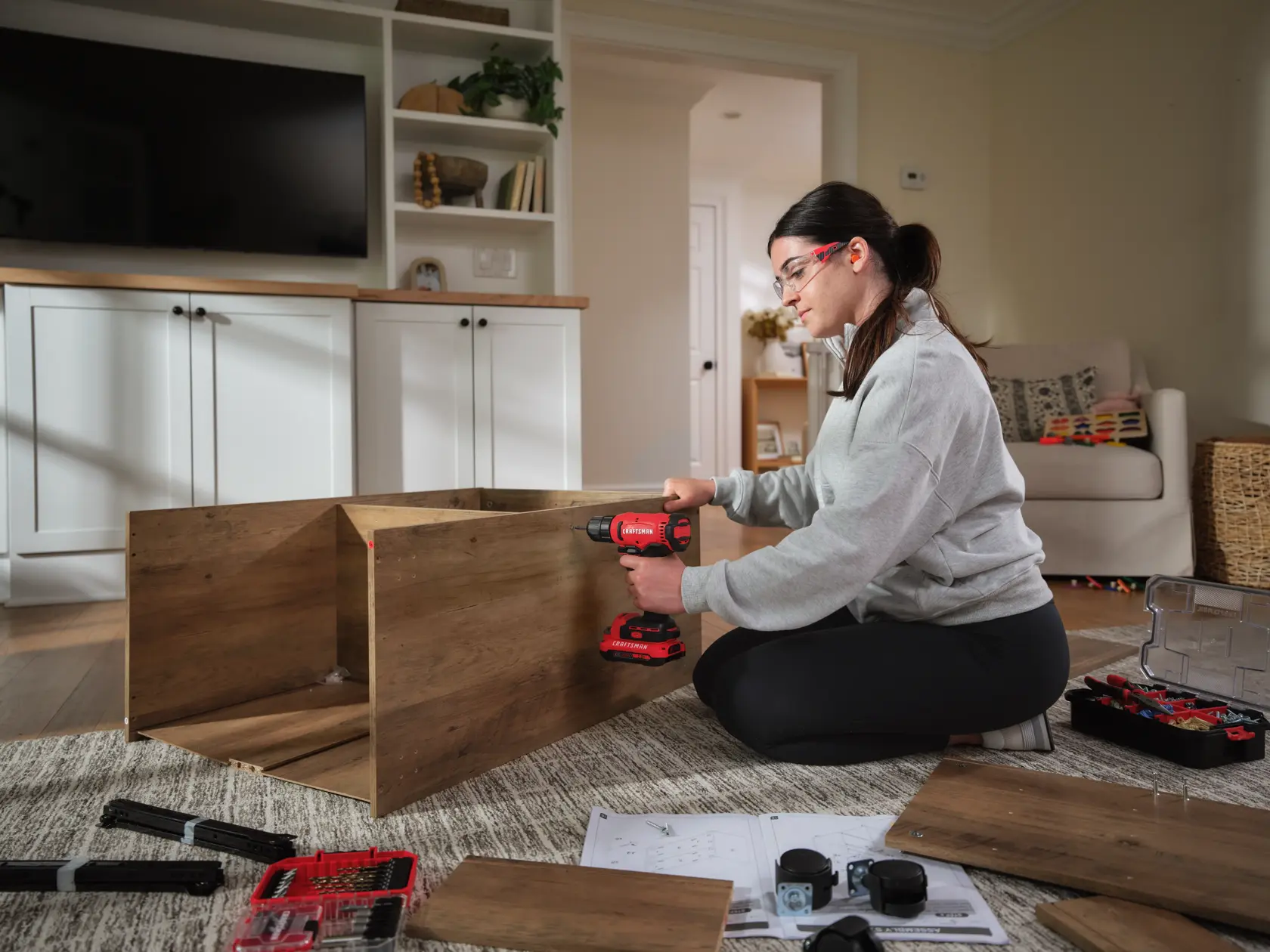 Person assembling wooden furniture on a carpeted floor using a red CRAFTSMAN cordless drill, with toolboxes, hardware, and instructions spread nearby in a living room setting.