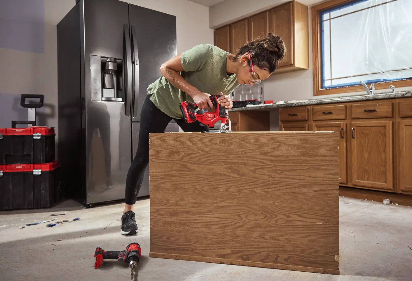 A person using a red CRAFTSMAN cordless drill to assemble or modify a wooden panel in a kitchen under renovation. The kitchen features wooden cabinets, a black refrigerator, and toolboxes in the background. Another red drill is lying on the floor.