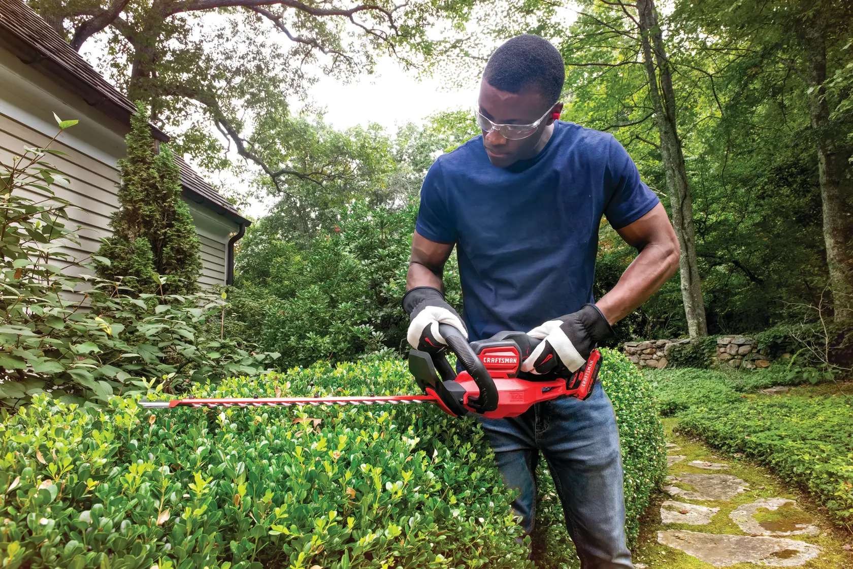 Photo of a person using a Craftsman hedge trimmer CMCHTS820D1 in a garden, trimming green bushes.