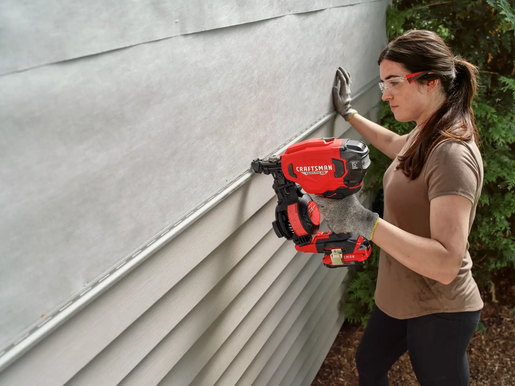 A person using a CRAFTSMAN cordless nail gun to install siding on an exterior wall. The individual is wearing gloves, a