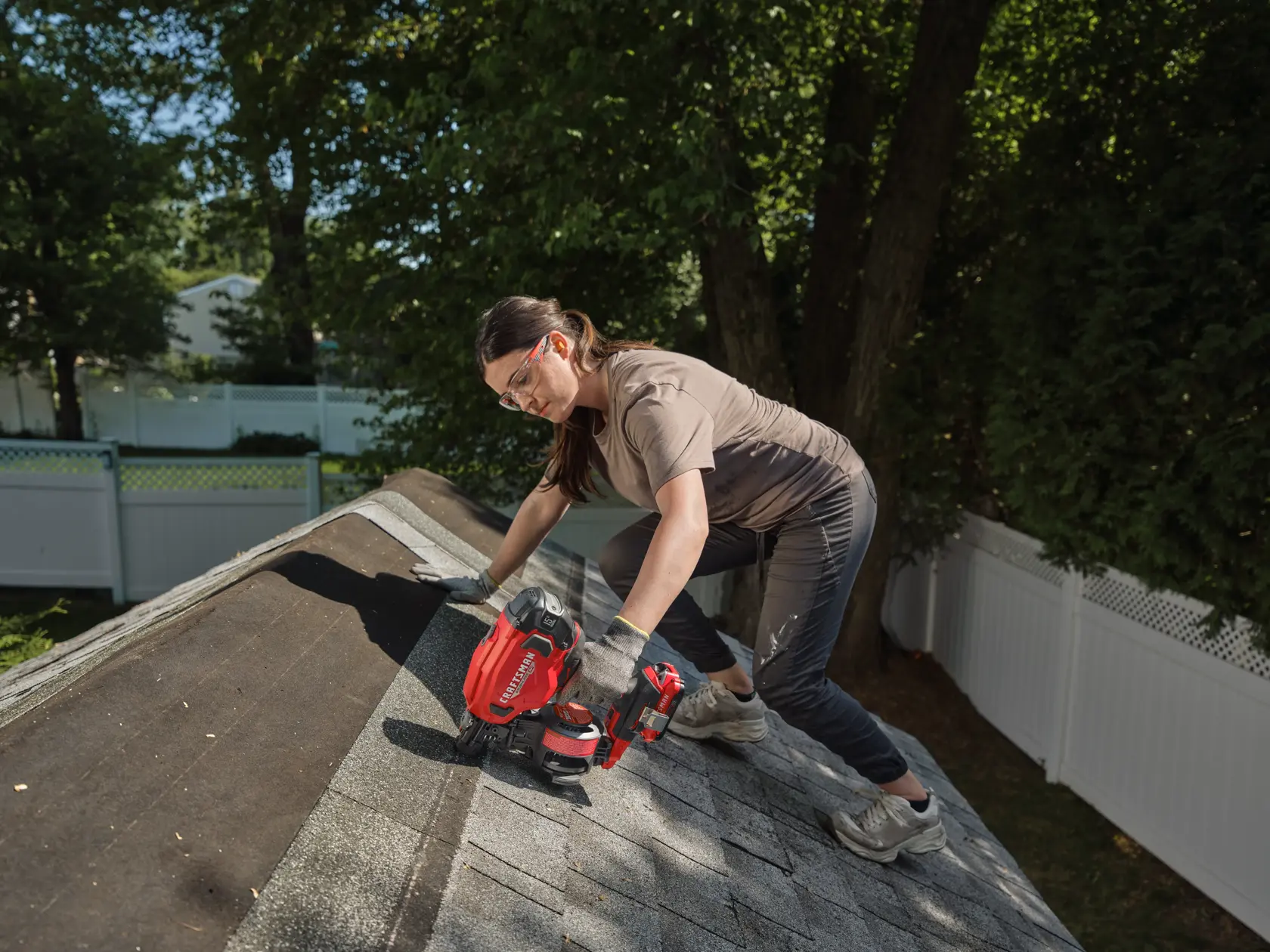 A person wearing safety gear is working on a rooftop using a red CRAFTSMAN branded cordless nailer. The person appears to be