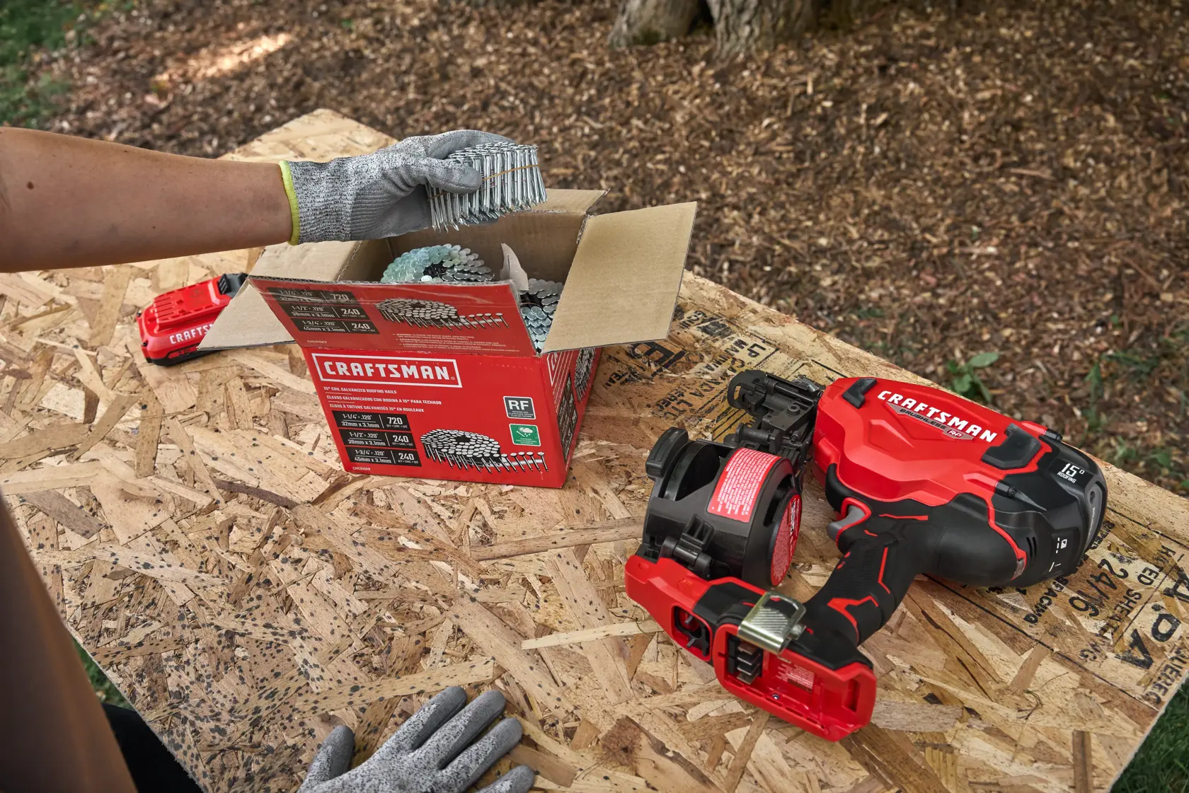 A close-up of a woodworking setup featuring a CRAFTSMAN box of coil nails, a battery pack, and a red and black CRAFTSMAN
