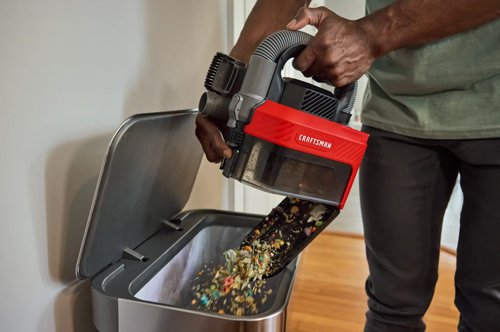 Person empties debris from a CRAFTSMAN vacuum into a trash bin in a home setting.