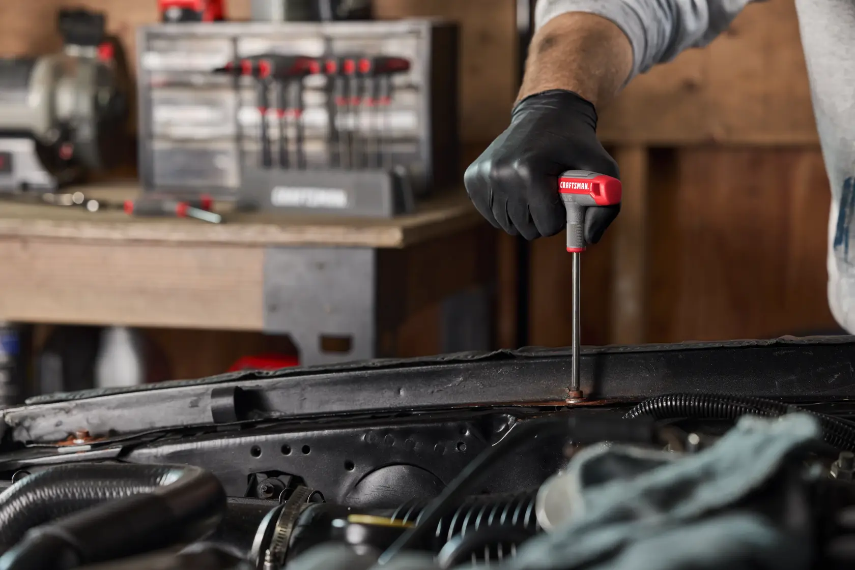 A person wearing black gloves is using a CRAFTSMAN T-handle hex key to work on a car engine. A set of T-handle hex keys is visible in the background on a workbench.