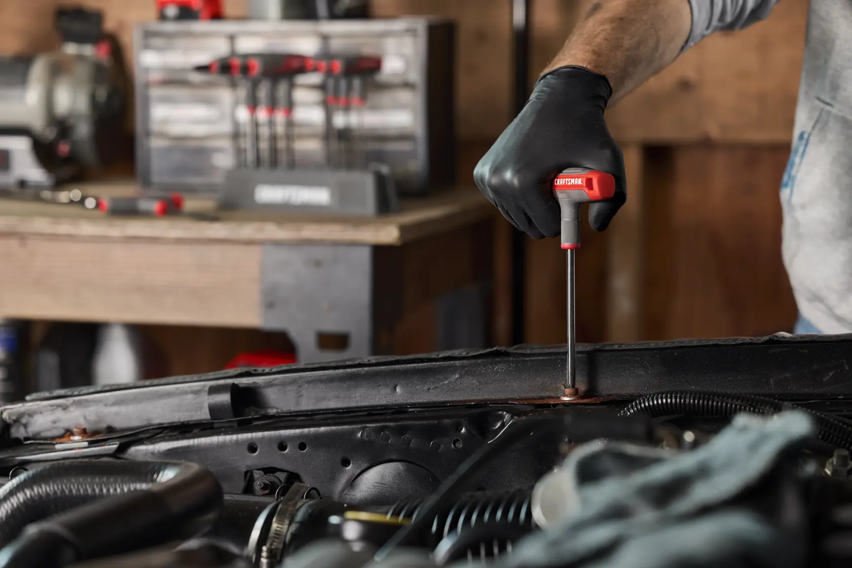 A person wearing a black glove uses a CRAFTSMAN T-handle screwdriver to work on the engine compartment of a vehicle. In the background, there is a set of tools and a workbench.