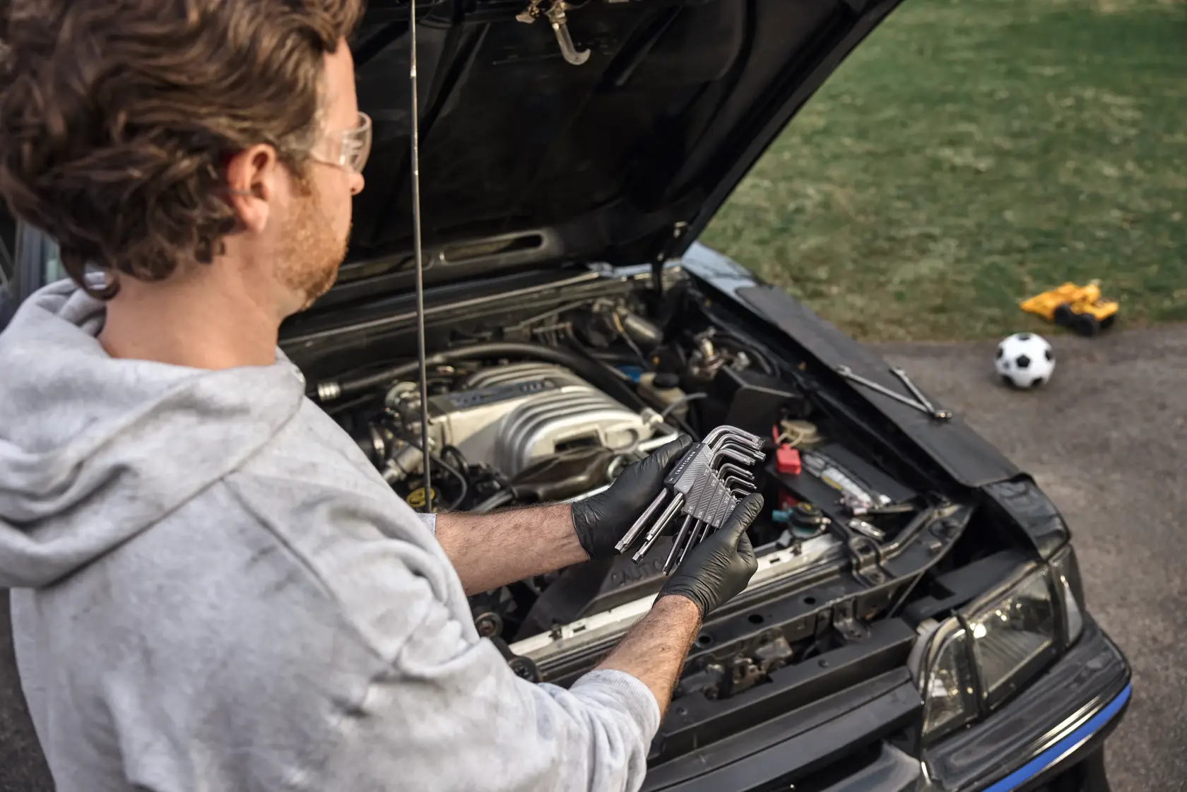 Person wearing gloves holds a set of hex keys while working on the engine bay of a car with the hood open. A soccer ball and toy construction vehicle are visible on the grass in the background.