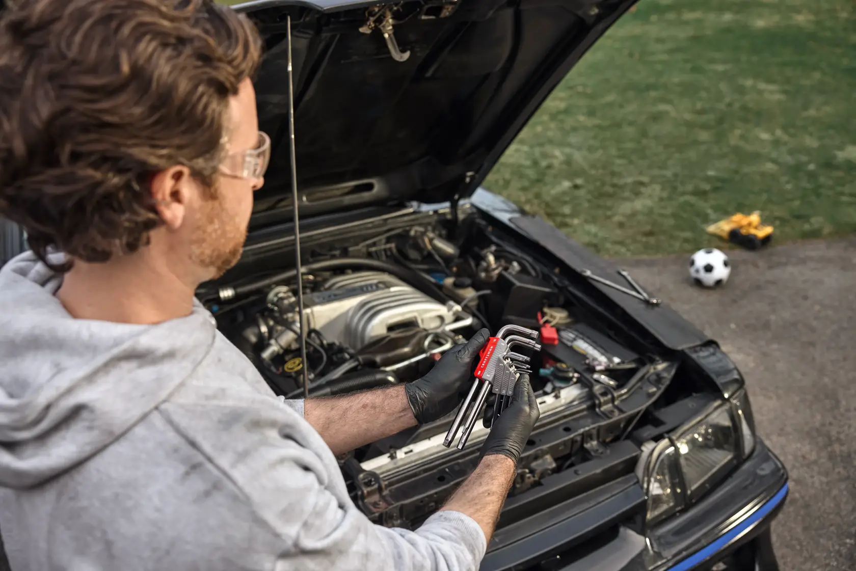 A person wearing safety glasses and gloves holds a set of CRAFTSMAN hex keys while working on a car engine with the hood open. The hex keys are in a red holder, and there is a soccer ball and toy truck in the background on the grass.