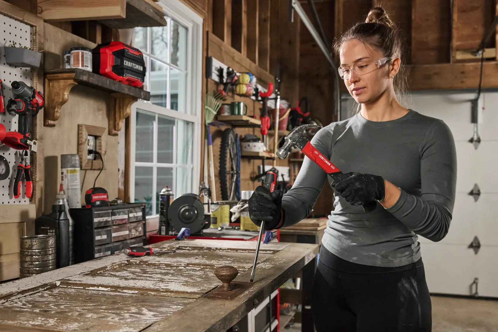 Person with face blurred wearing black gloves and a grey shirt uses a CRAFTSMAN screwdriver and hammer while working on a wooden surface in a workshop with tools and equipment visible in the background.