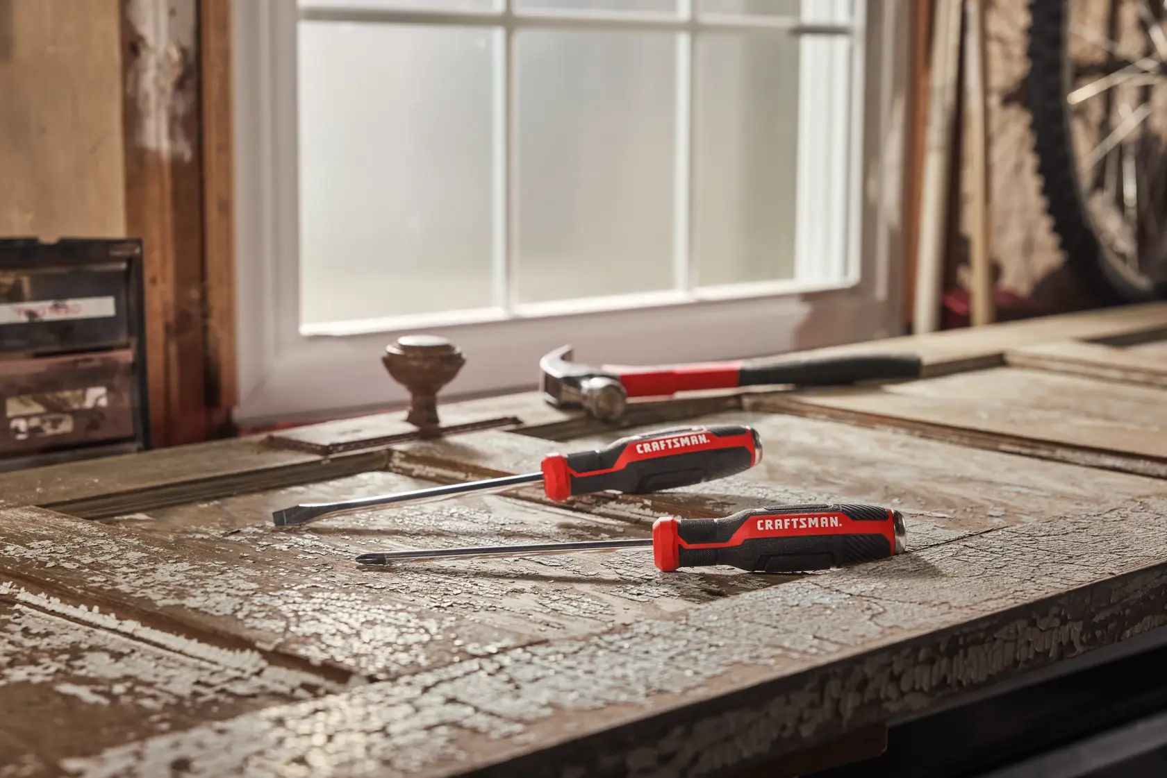 Three CRAFTSMAN hand tools, including two screwdrivers and a hammer, are placed on a worn and textured wooden surface near a window in a workshop setting.