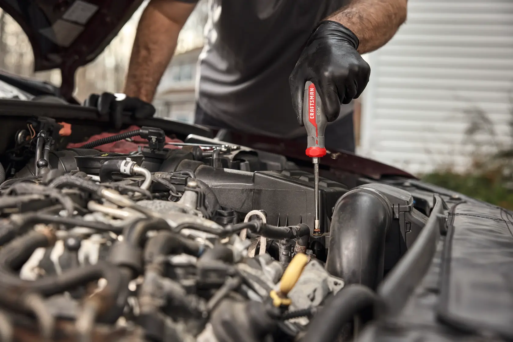 A person wearing black gloves is using a CRAFTSMAN screwdriver to work on the engine of a car.