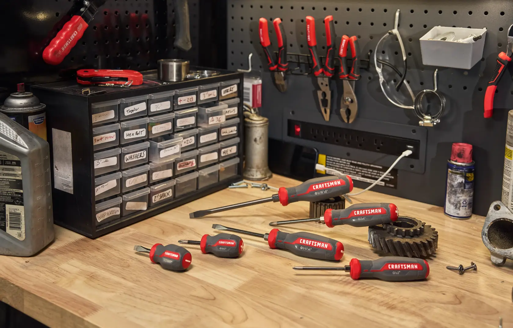 A set of CRAFTSMAN screwdrivers with red and black handles arranged on a wooden workbench in a workshop. Behind the screwdrivers, there are storage drawers labeled with parts, pliers hanging on a pegboard, and various workshop supplies.