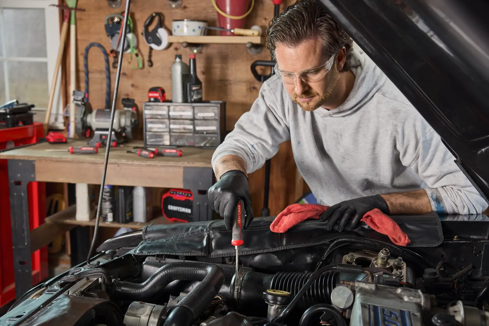 A person wearing gloves is using a CRAFTSMAN screwdriver to work under the hood of a car in a garage workshop. The person's face is blurred. Various tools and red cloths are visible on the car and the workbench in the background.