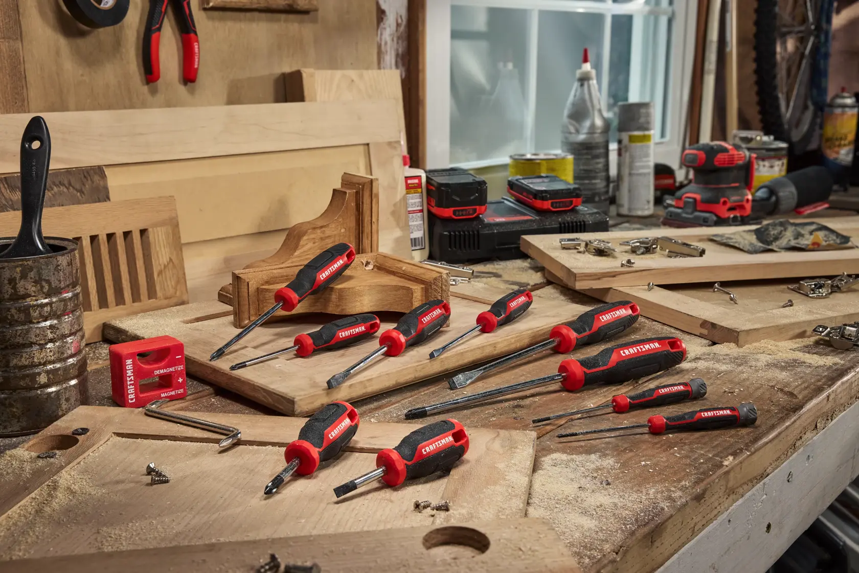 A set of CRAFTSMAN screwdrivers with red and black handles arranged on a woodworking bench, surrounded by wooden boards, screws, and various workshop tools.