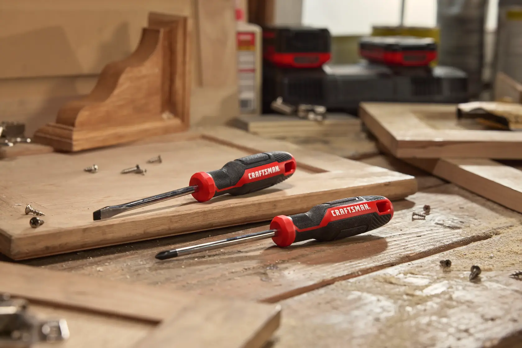 Two CRAFTSMAN screwdrivers with black and red handles are placed on a wooden workbench surrounded by wood pieces and metal screws. The workspace appears to be a carpentry or workshop setting.