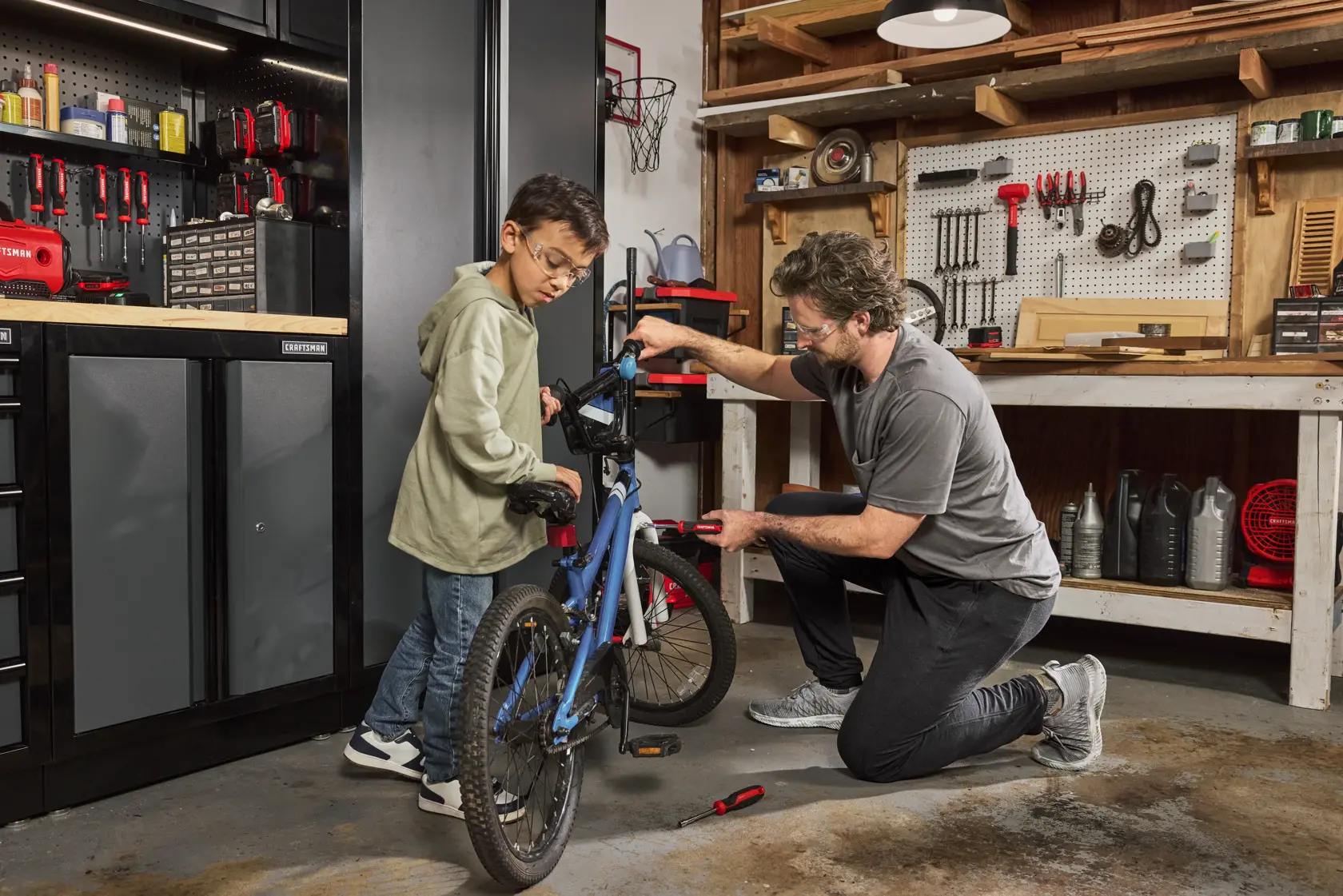 A person kneeling in a workshop uses a hand tool to repair a blue bicycle while another person stands nearby. The workshop has organized storage cabinets, pegboard tool storage, and shelves with various tools and supplies.