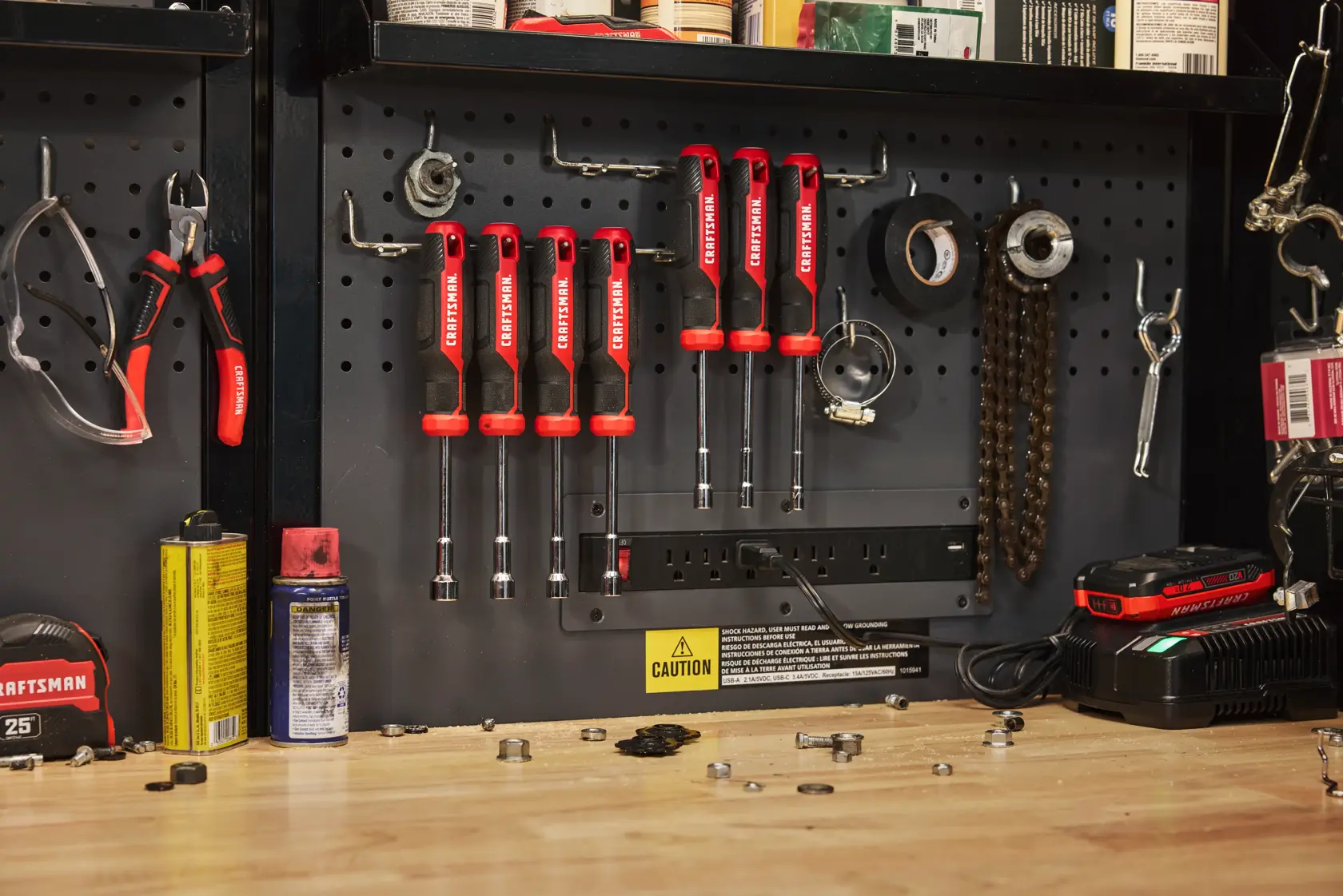 A workshop scene showing a pegboard with various tools including a set of CRAFTSMAN nut drivers with red handles, pliers, safety glasses, tape, a chain, lubricant cans, a tape measure, scattered bolts, nuts, and a battery charger on a wooden workbench.