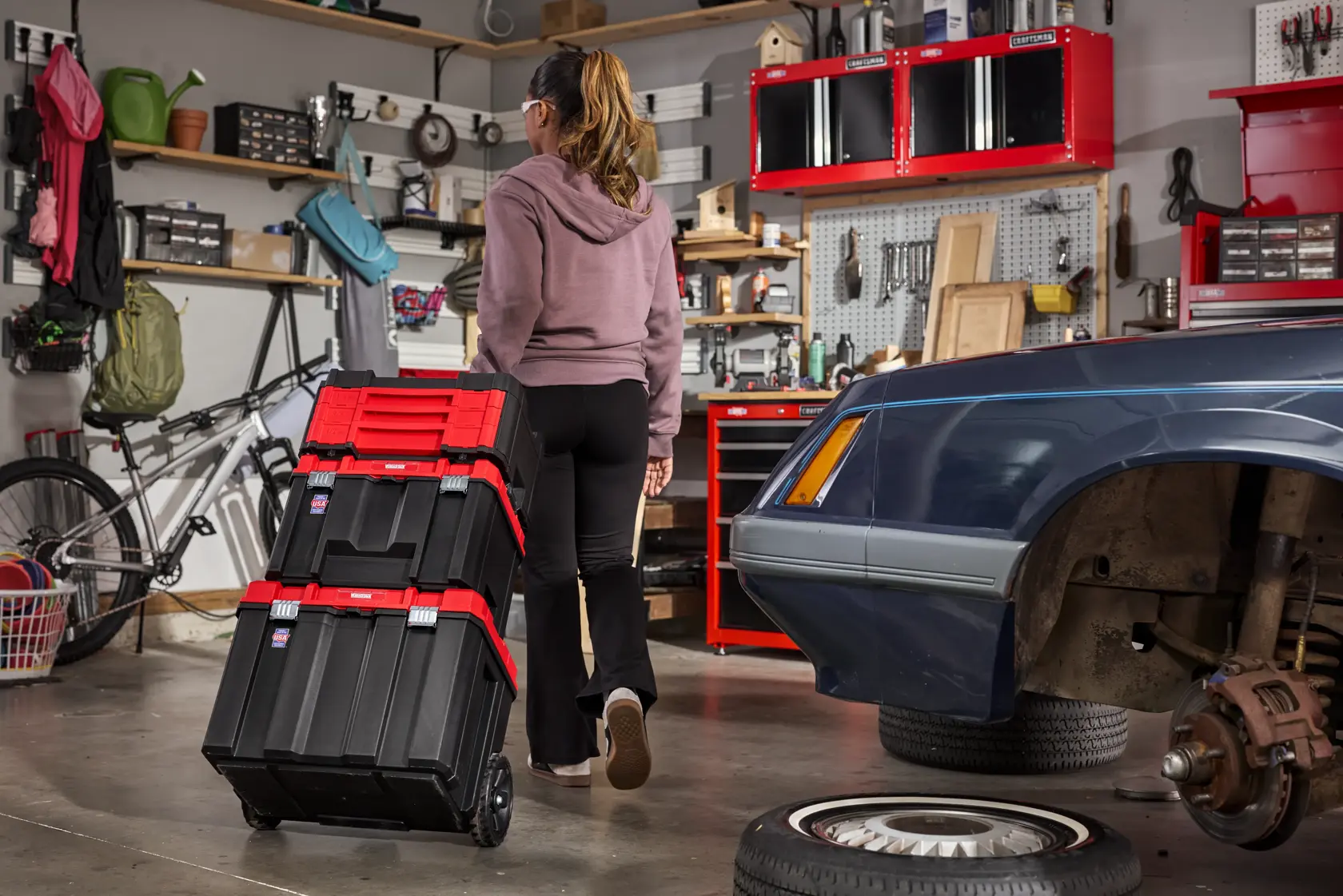 A person is pulling a stack of black and red rolling toolboxes in a garage or workshop. The space is organized with shelves, a bicycle, and various tools. A car is partially visible with its wheel removed, and other automotive tools are nearby.