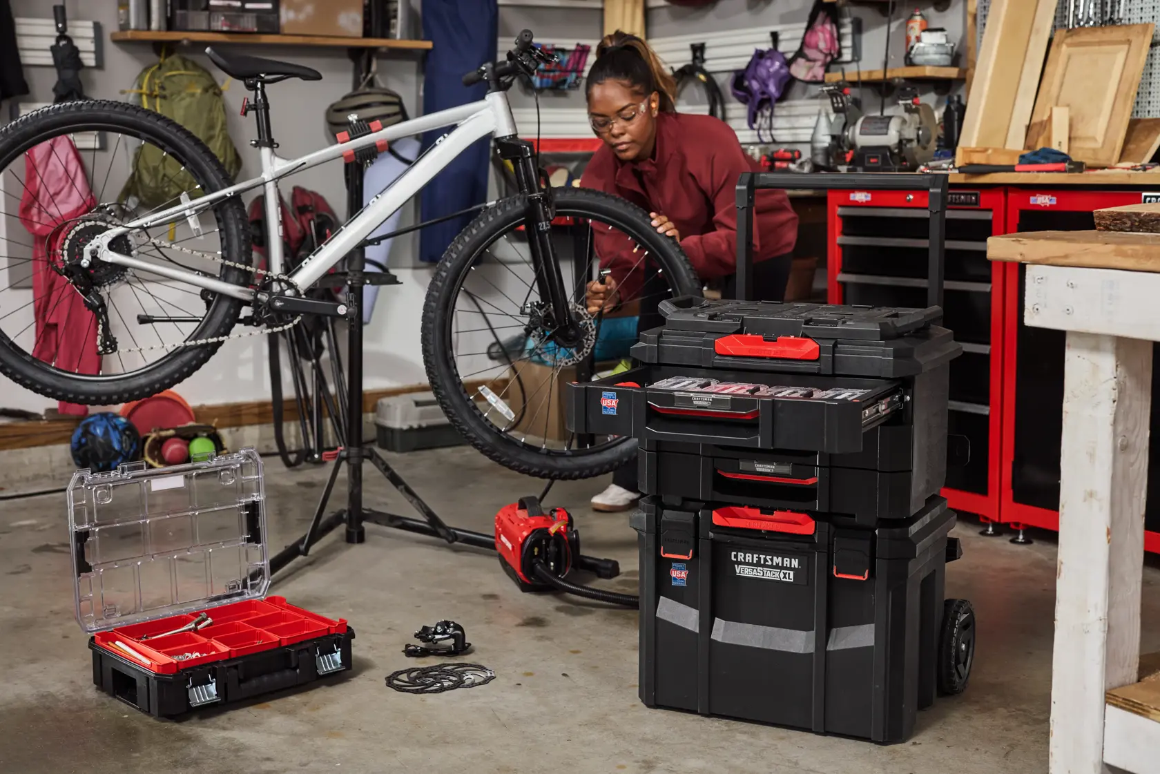 A garage workspace featuring a white bicycle mounted on a bike repair stand, surrounded by various tools and toolboxes including a stackable Craftsman tool storage system and an open tool case. Shelves and red cabinets filled with equipment and supplies are visible in the background.