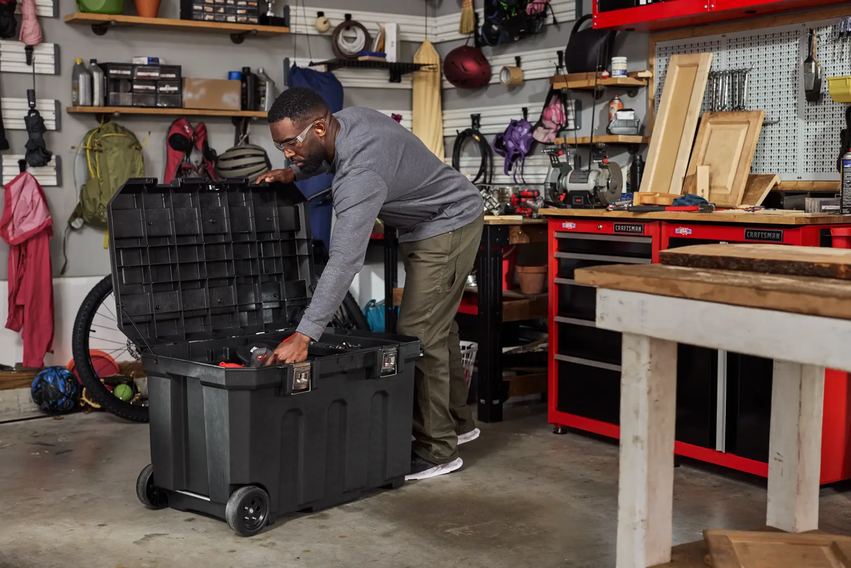 A person is standing in a workshop or garage, using a large black rolling storage chest with an open lid. The workspace is organized with shelves, tools, tool chests, and various items like backpacks, wood pieces, and other equipment visible in the background.