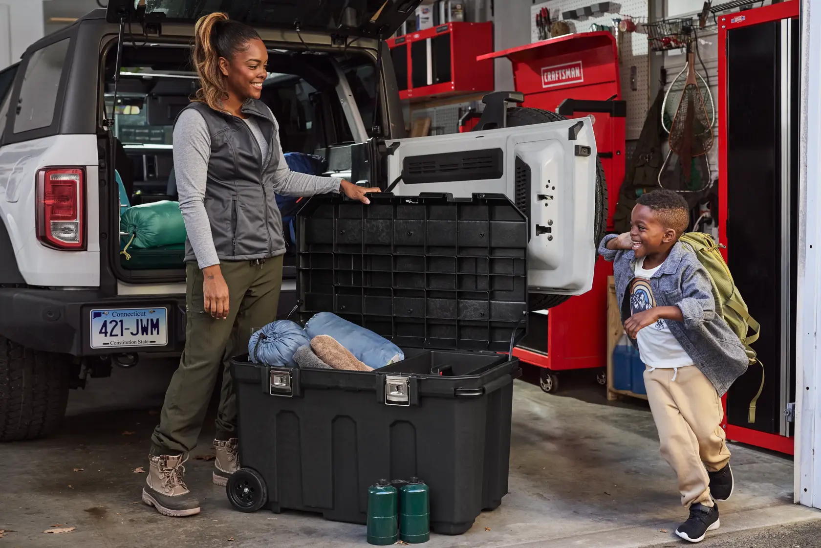 A large black storage container with an open lid is placed on the ground next to a vehicle with its rear door open. There are camping items such as sleeping bags inside the container, and two green fuel canisters placed in front of it. The background shows shelves with tools and outdoor equipment in a garage.