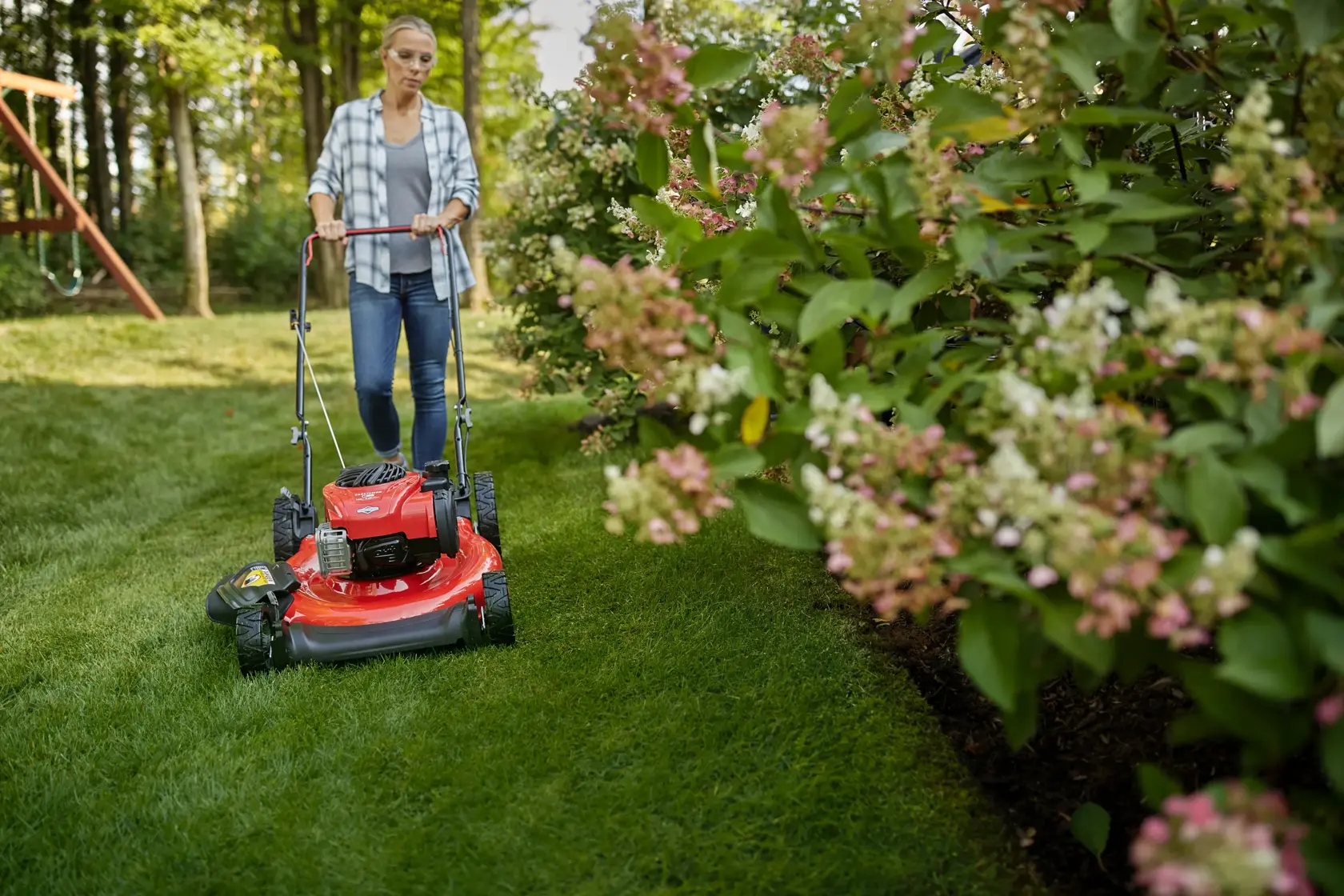 CRAFTSMAN M100 140cc Push Mower mowing near flower bed with bushes wearing jeans and plaid button up shirt