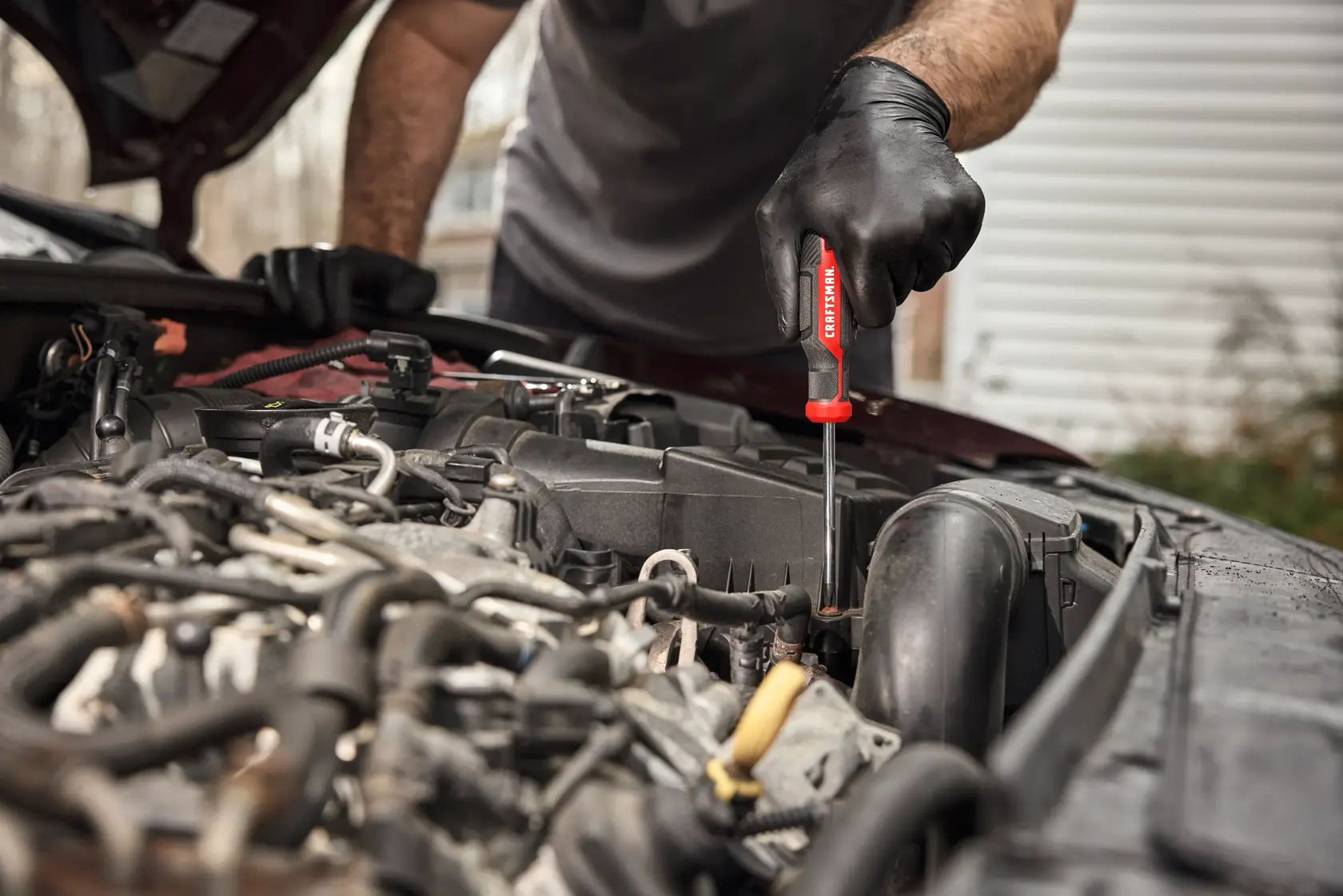 A person wearing black gloves uses a red-handled CRAFTSMAN screwdriver to work on the engine of a car with the hood open.