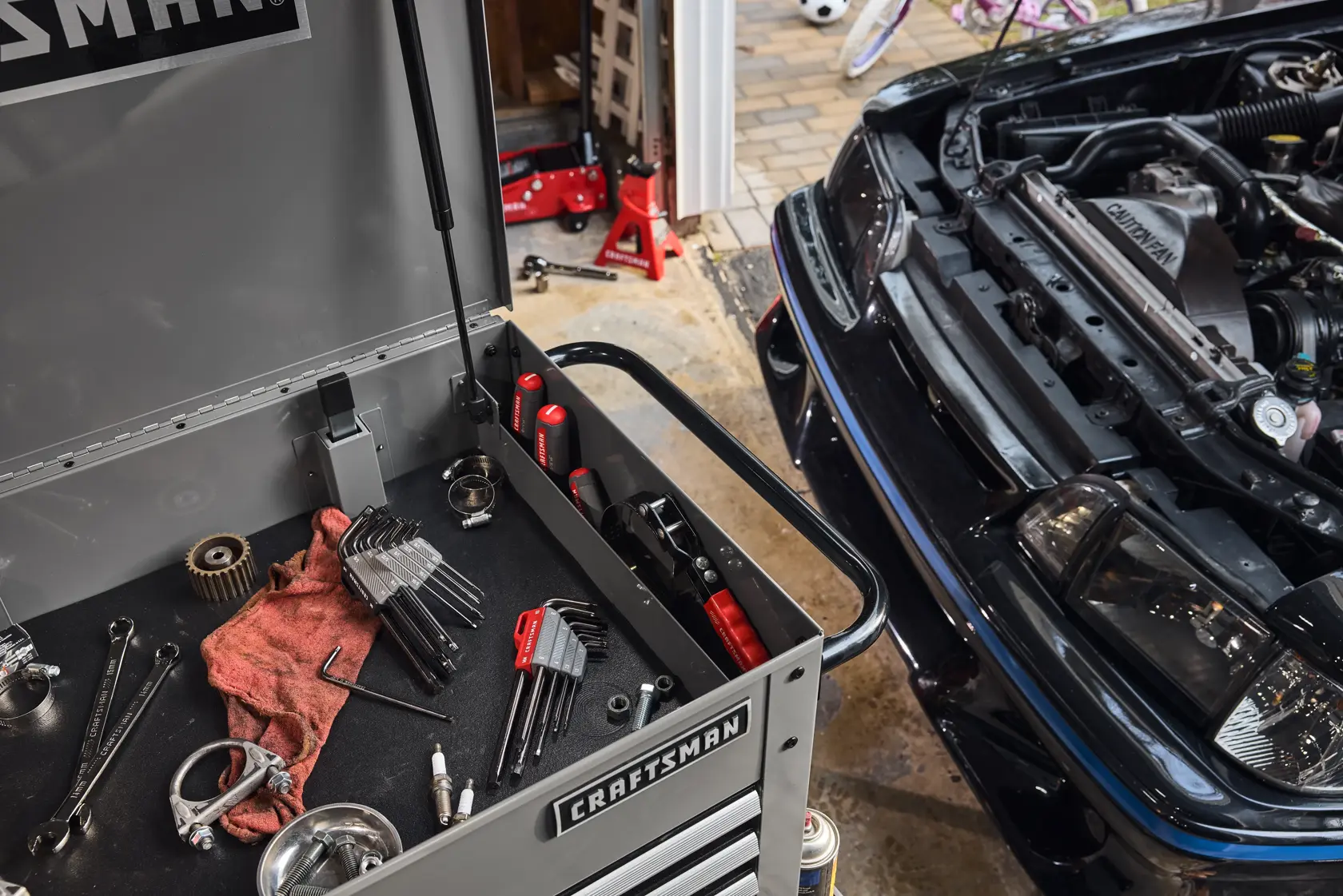 A CRAFTSMAN tool chest with various automotive tools and accessories is open next to a black car with its hood lifted, inside a garage. The image shows hex keys, wrenches, spark plugs, a red cloth, and other tools on the tool chest. The car engine and surrounding area are visible, with additional tools and equipment in the background.