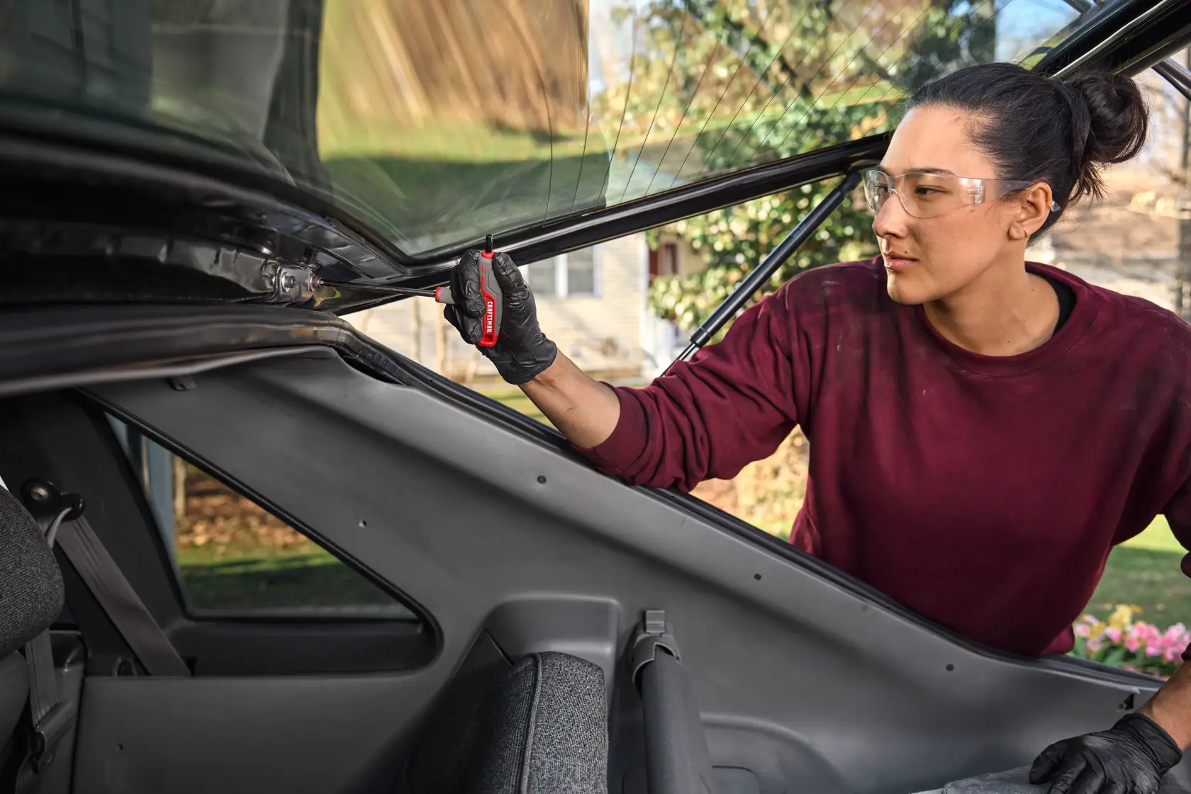 A person wearing black gloves and a maroon shirt is using a CRAFTSMAN tool to work on the inside part of a car's rear hatch. The person's face is blurred, and the scene is set outdoors.