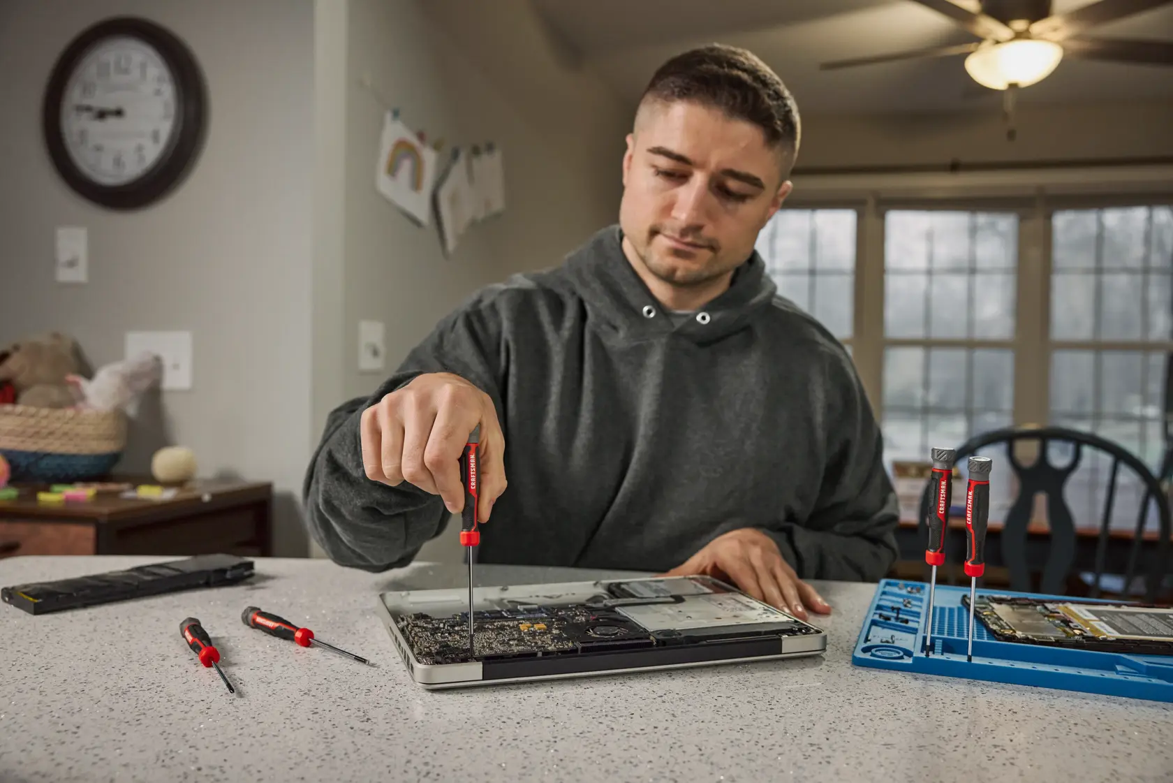 A person using a CRAFTSMAN precision screwdriver to repair a laptop on a kitchen counter, with additional screwdrivers and small tools nearby. The person's face is blurred.