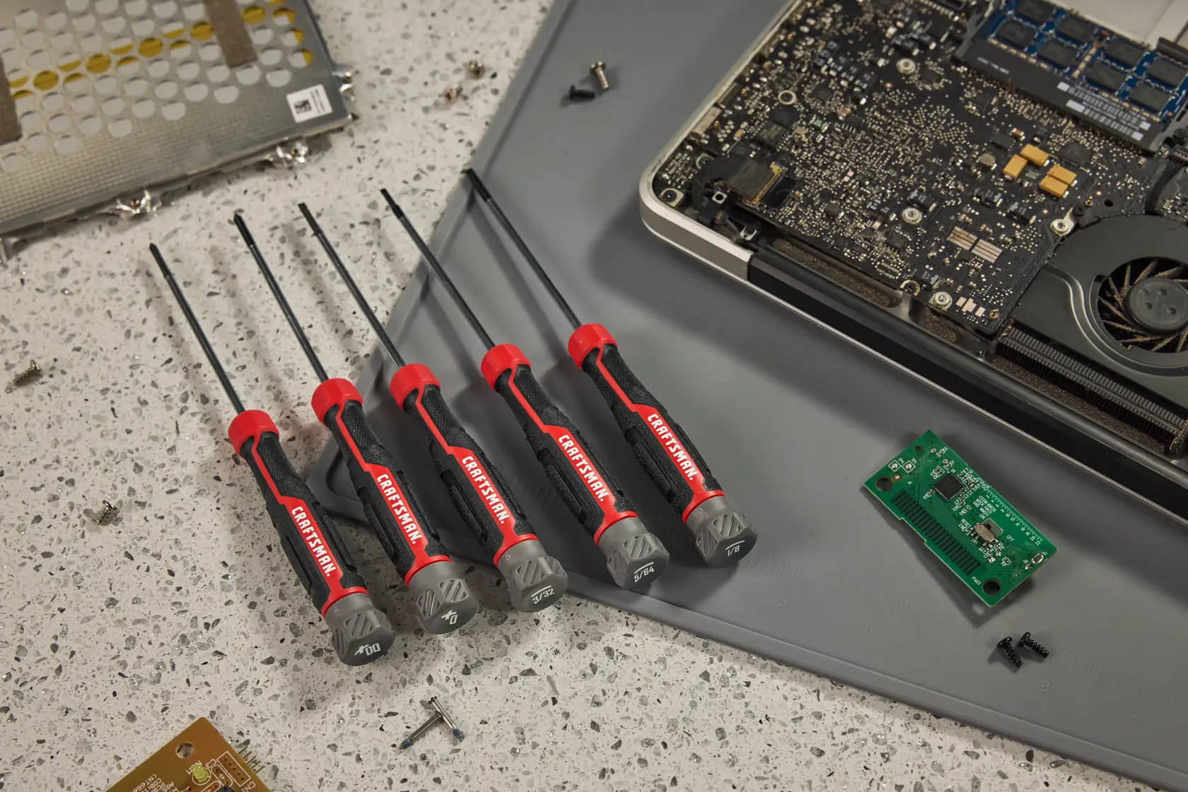 Five CRAFTSMAN precision screwdrivers with black and red handles are arranged on a work surface next to an open electronic device, circuit boards, and small screws.