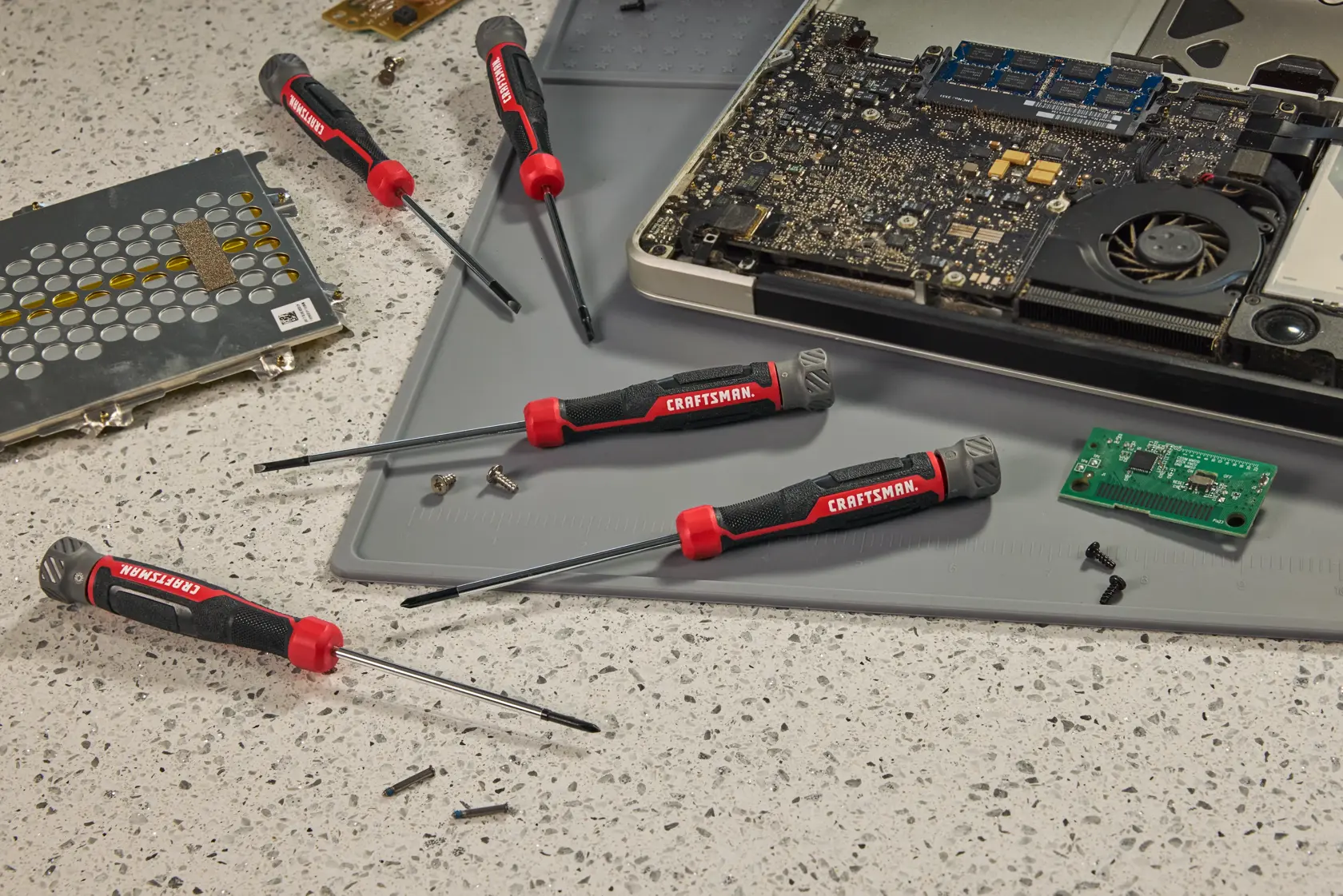 A set of CRAFTSMAN precision screwdrivers are shown on a work surface with electronic components including a partially disassembled laptop, a circuit board, screws, and a gray mat.