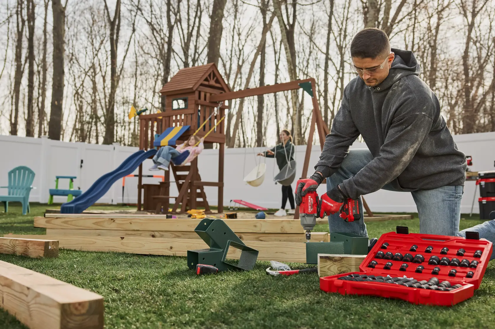 A person is kneeling on the grass using power tools to assemble wooden playground equipment. A red tool kit with various sockets and tools is open in the foreground. In the background, a child is on a swing and another person is standing nearby. The setting is an outdoor yard with a play structure and a white fence.